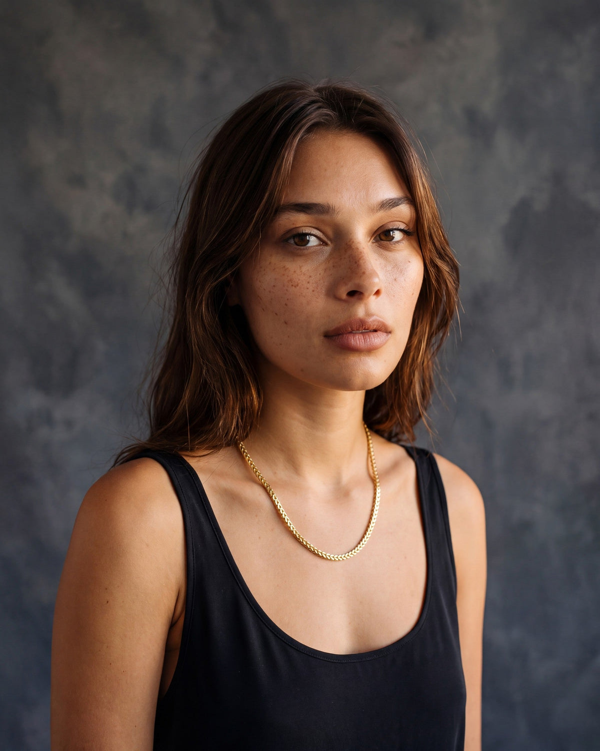 Woman wearing a 14K yellow gold diamond-cut Franco chain with a black tank top against a charcoal painted canvas backdrop