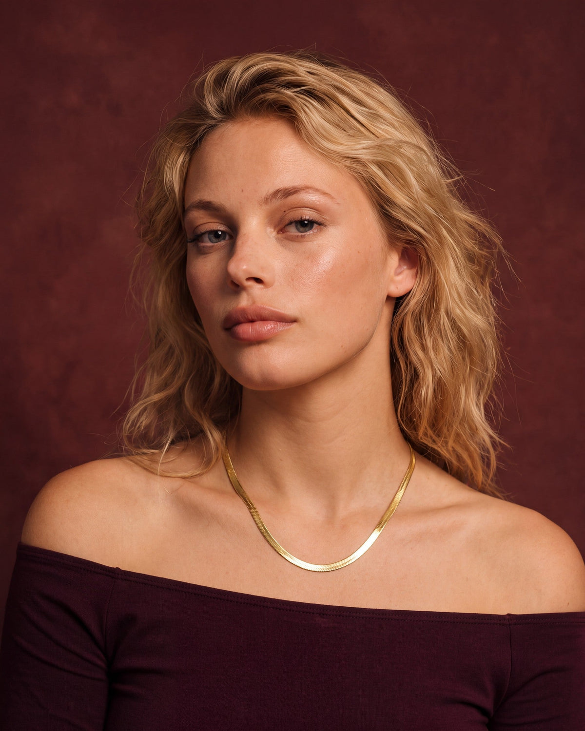 Woman wearing a 14K yellow gold Super Flex herringbone chain with an off-shoulder wine top against a wine-red painted canvas backdrop