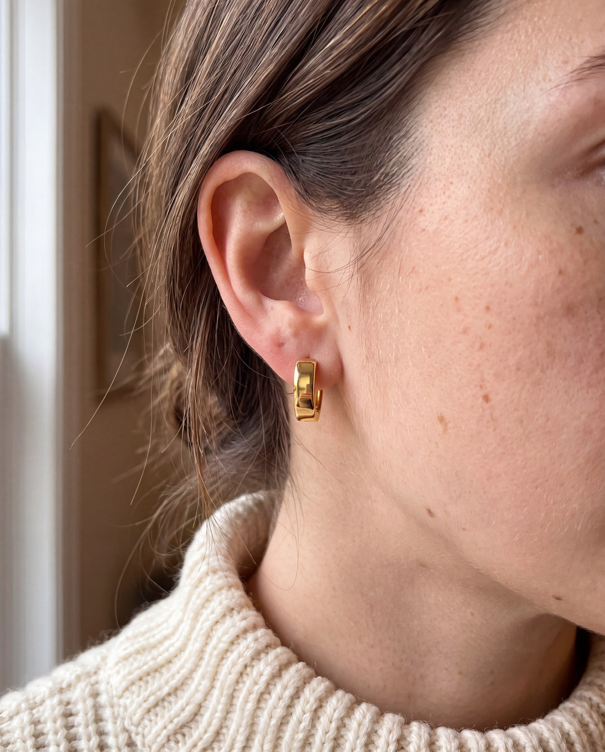 Close-up of a 14K yellow gold huggie earring worn on the earlobe, cream knit sweater, window light