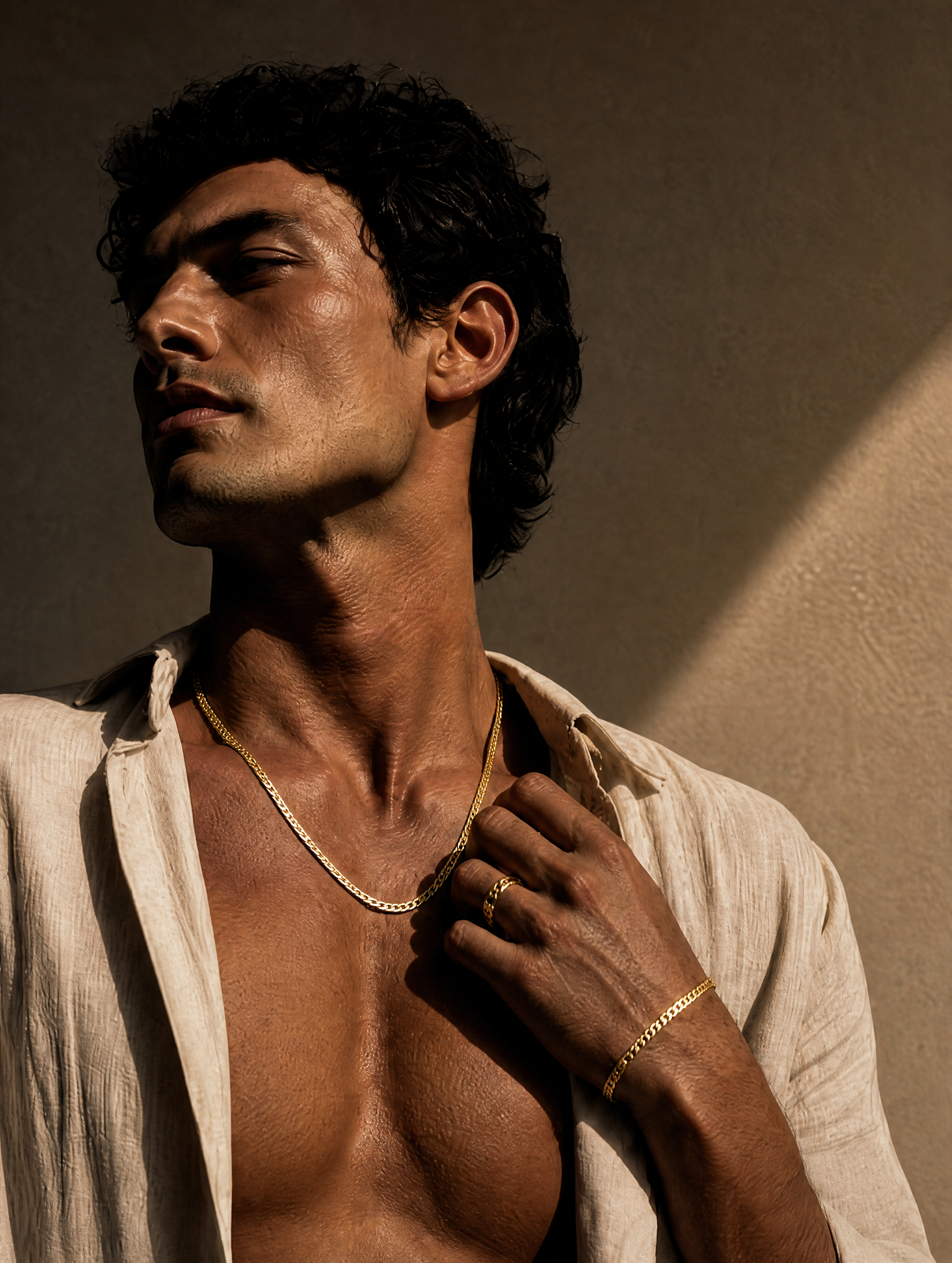 A man wearing a delicate gold chain necklace and matching gold bracelet, styled with an open white linen shirt against a textured beige wall.